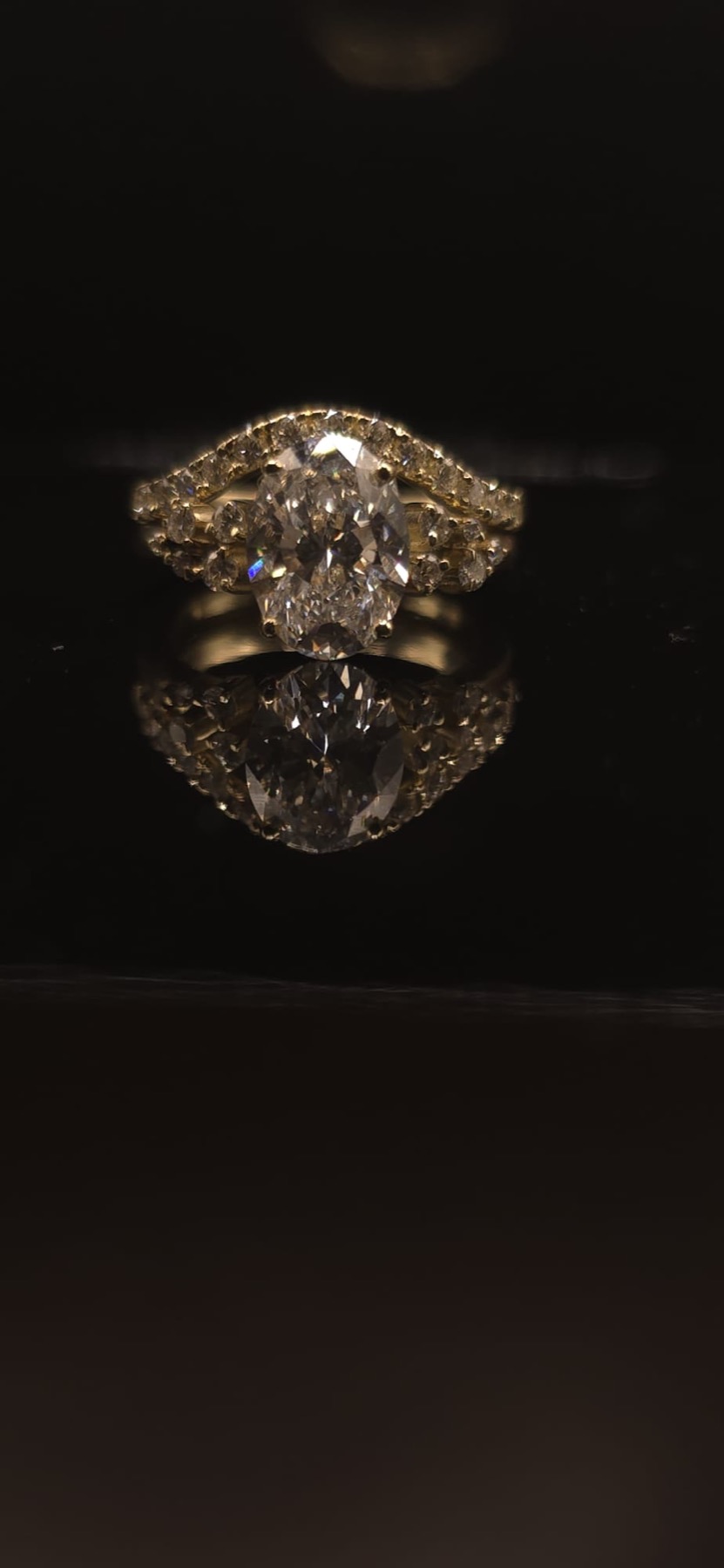 Oval diamond engagement ring with pavé band, reflected on a dark surface